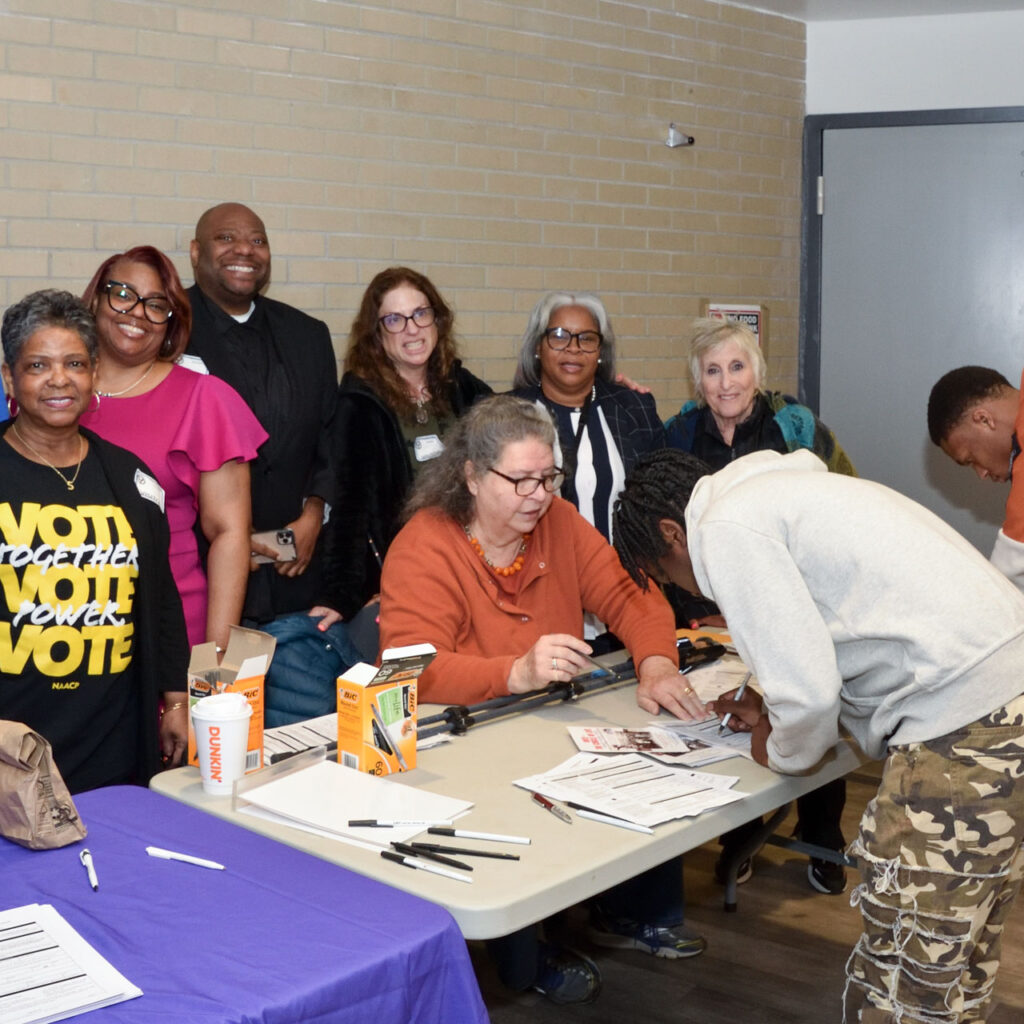 A crowd of people stands behind a table. Two young men are bent over the table signing papers.
