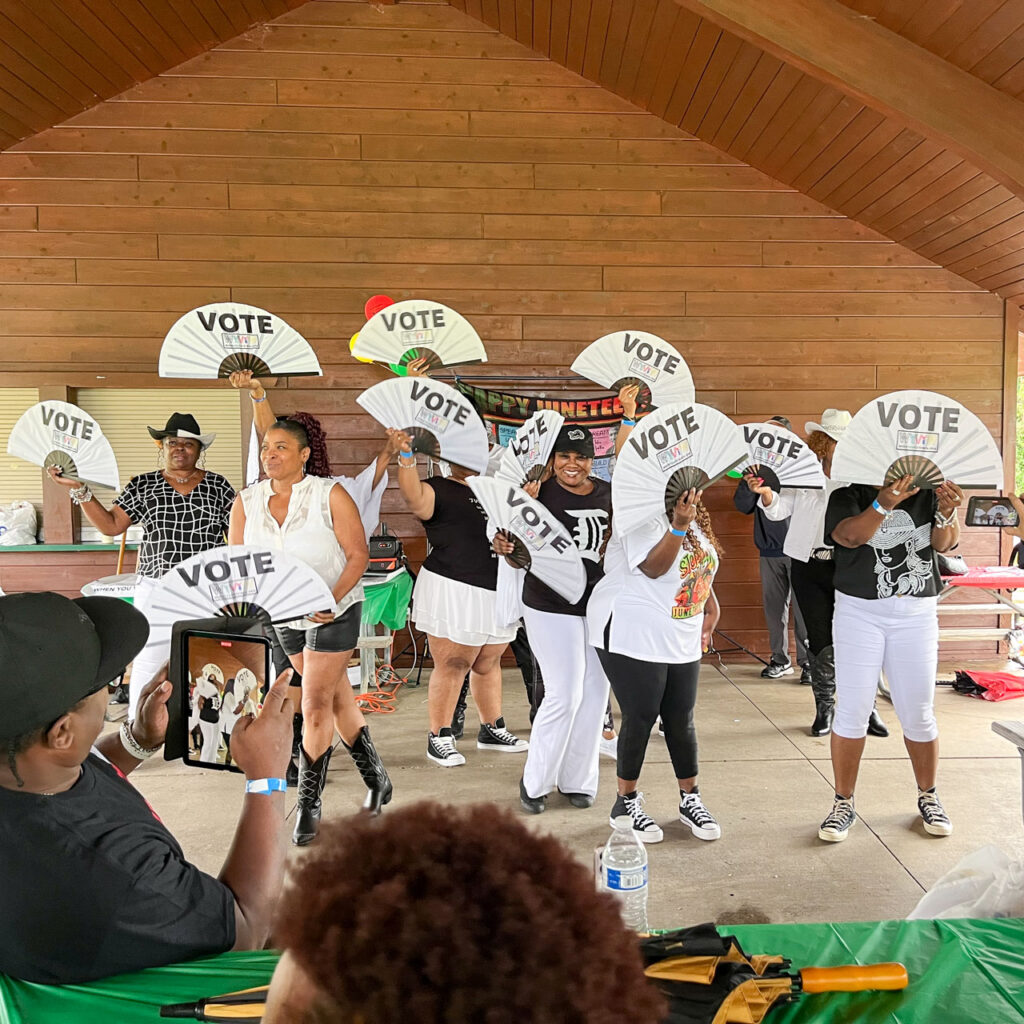 A group of people in a shelter waving fans printed with the word, vote.