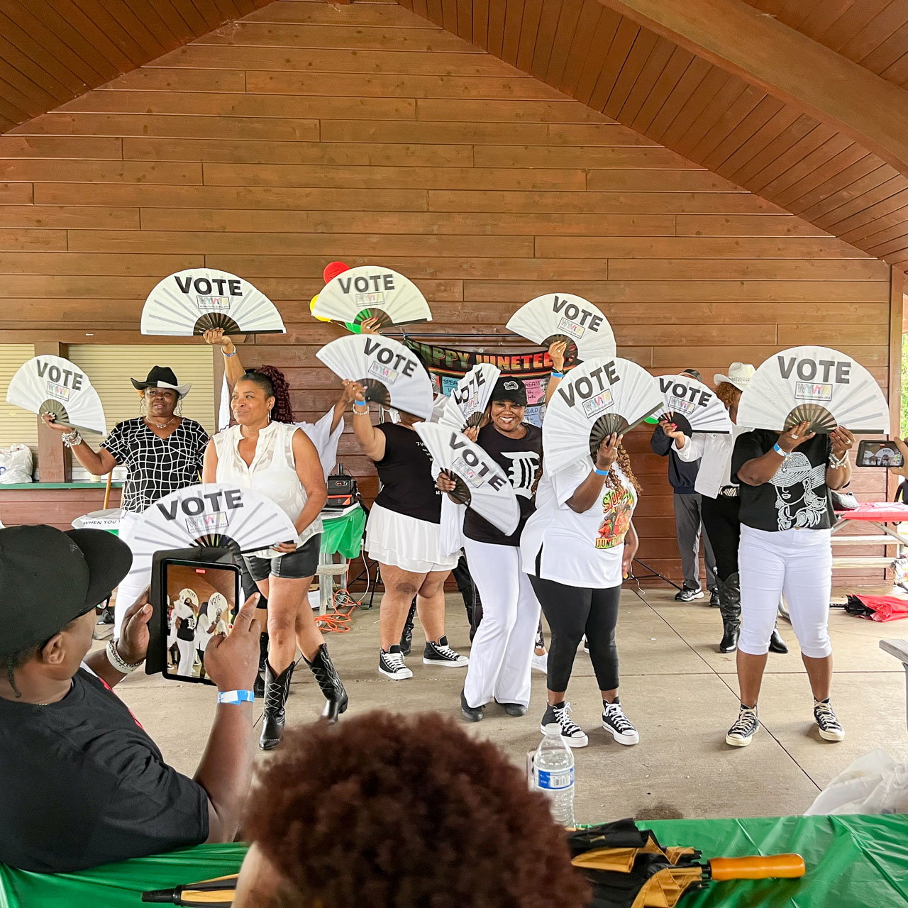 A group of people in a shelter waving fans printed with the word, vote.